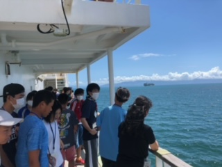 たくさんの参加者たちが船のデッキに出て、青空と海を眺めながら説明を受けている様子の写真