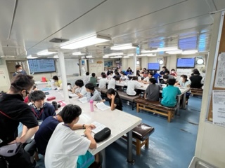 白く広い船内の研修室で、多くの参加者が長テーブルを囲んで座り、真剣な表情で配布された資料に目を通している様子の写真