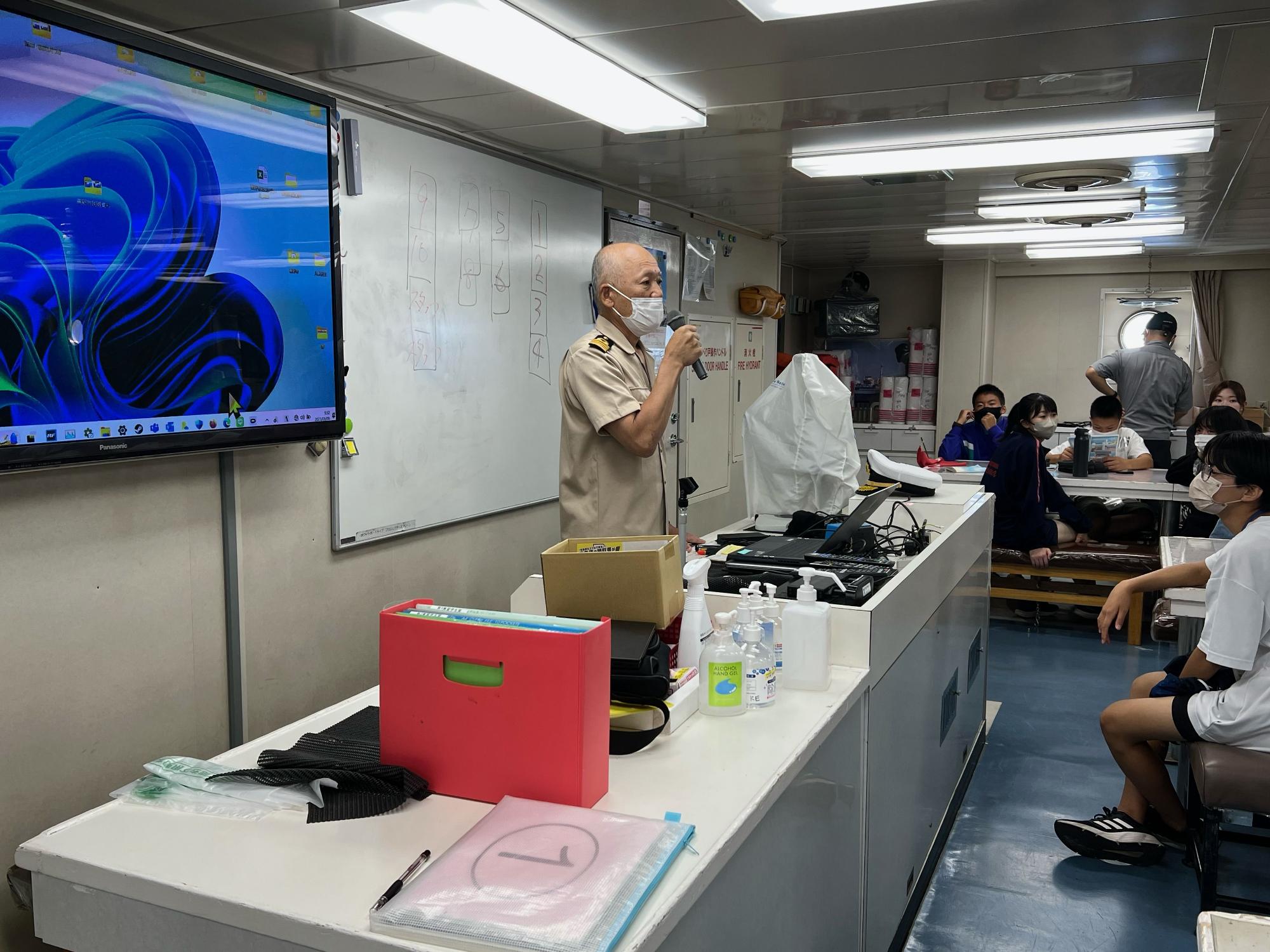 マイクを持った制服姿の船長が、白板やディスプレイが設置された船内の研修室で、テーブルに座る複数の参加者に向かって話している様子の写真