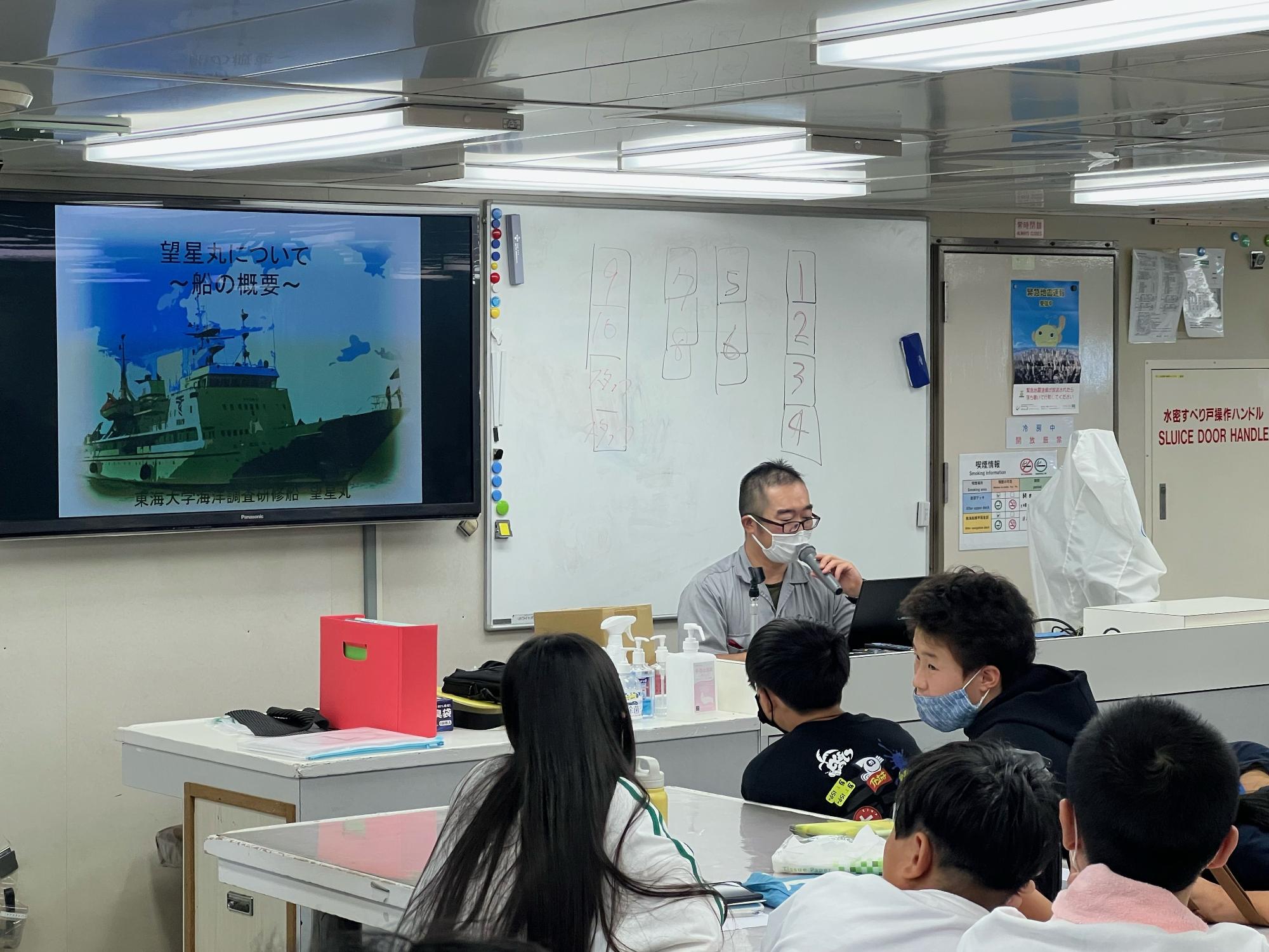 白いボードとスクリーンがある船内の研修室で、スクリーンに「望星丸について」の文字と船のイラストが表示され、マイクを持った講師が参加者に向かって話している様子の写真