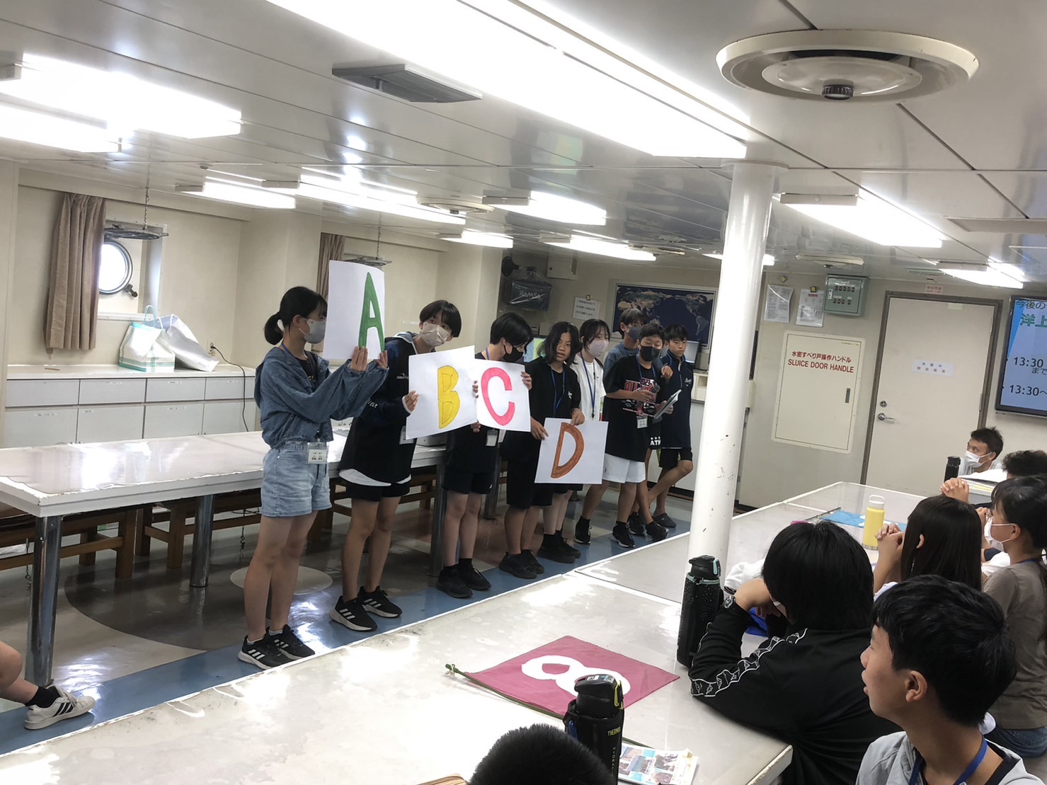 船内の部屋で、子どもたちががそれぞれAからDの文字が書かれた白い紙を持って並んで立ち、座っている子どもたちに出し物を披露している様子の写真