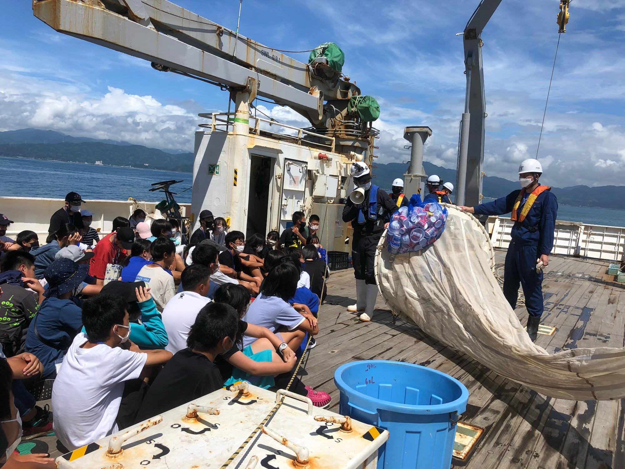 たくさんの子どもたちが船の甲板に座り、作業着とヘルメットを着用した乗組員からプラスチックごみなどが入った網や大きな網を見ながら説明を受けている写真