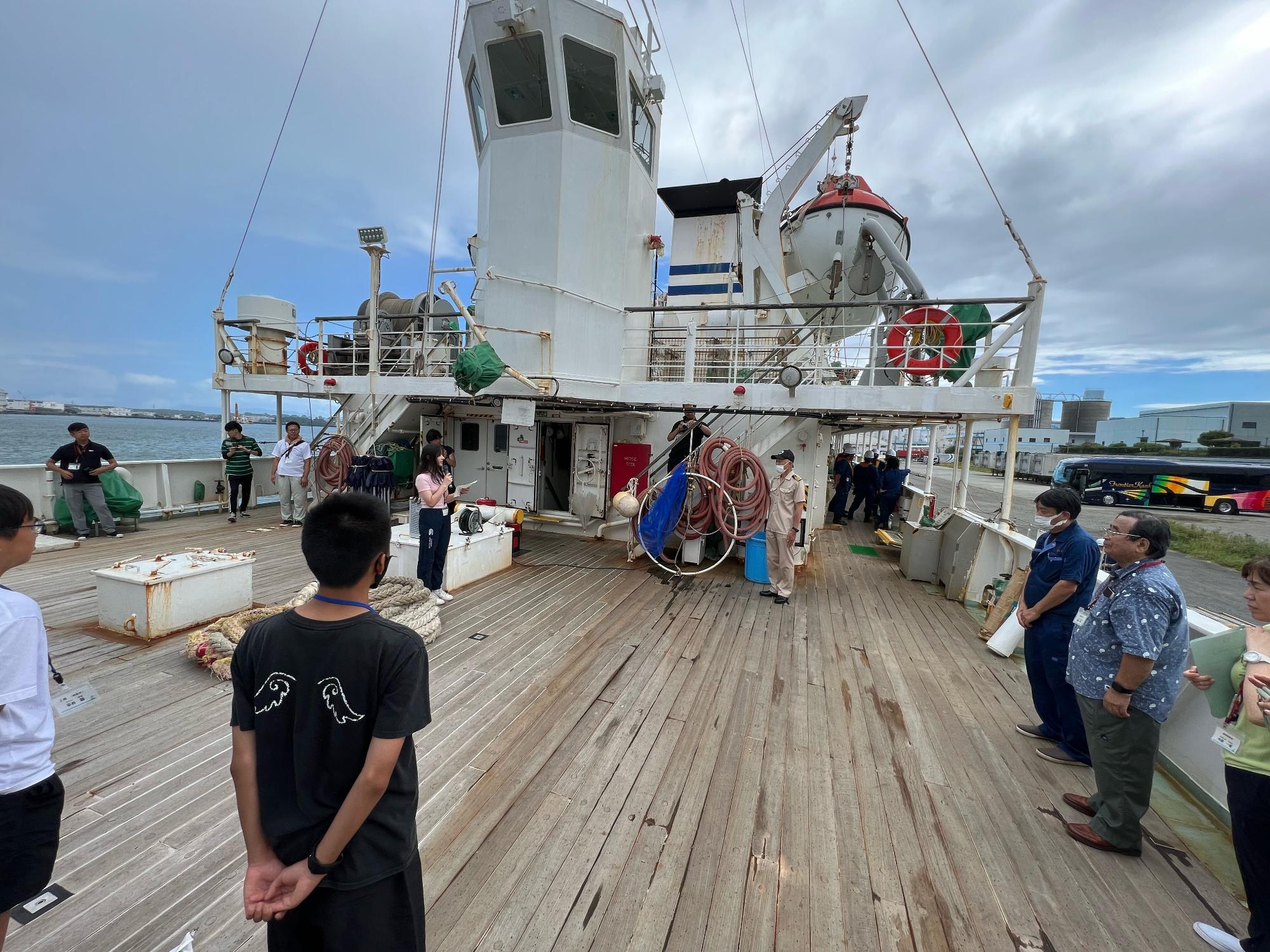 船のデッキで船長の男性と向かい合って立った参加者の女子生徒がマイクを持って挨拶をしており、周りで参加者や関係者が見守っている様子の写真