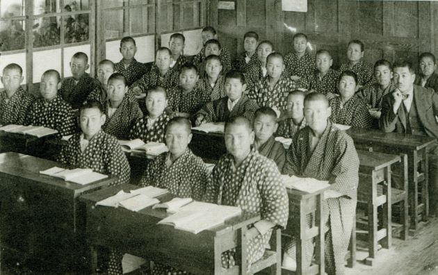 教室の席に大勢の和服を着用した坊主頭の男子生徒と風名の男性教師が座ってカメラの方を向いている白黒の集合写真