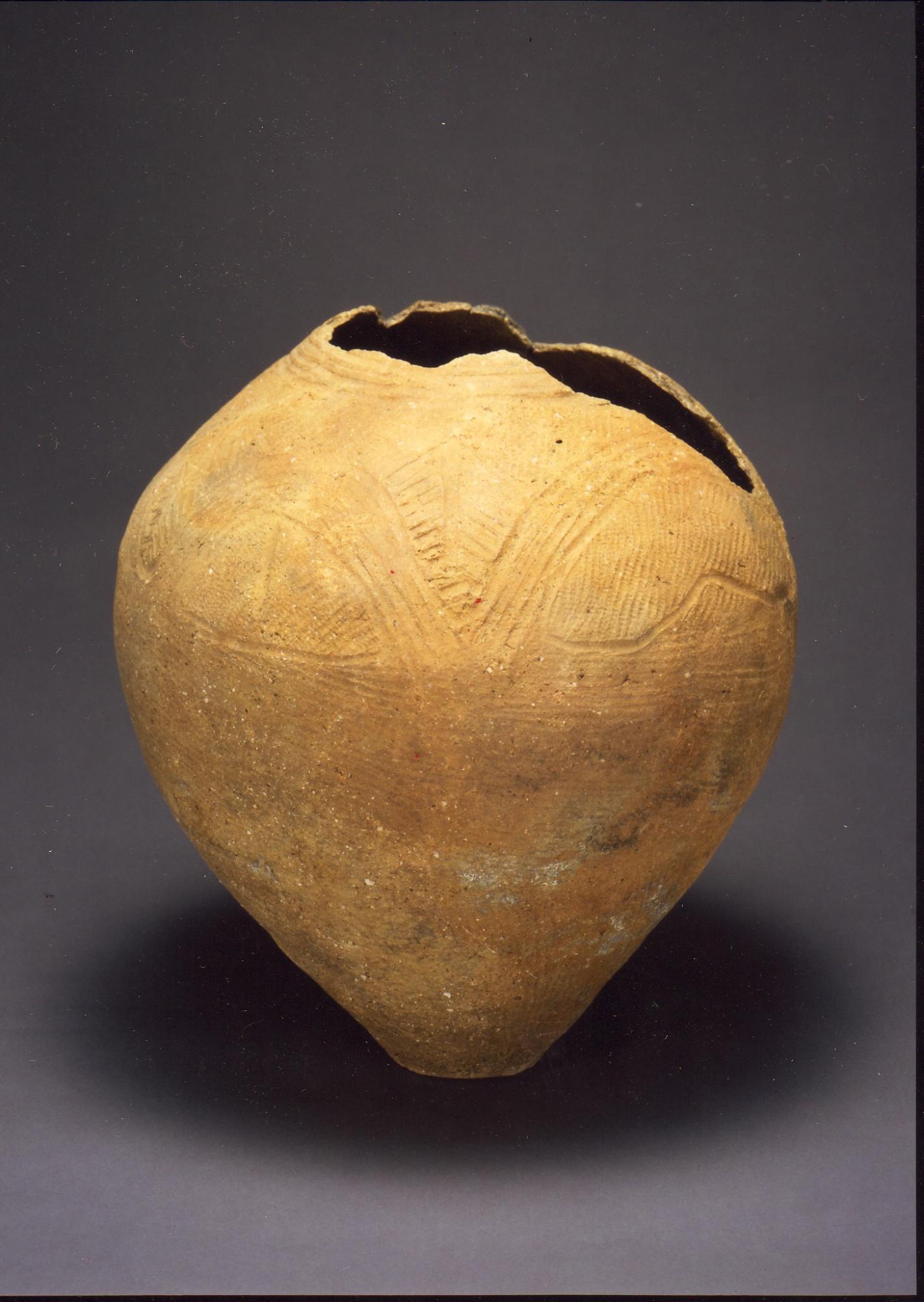 全体が丸みを帯び、底にかけて細くなった壷形で、上部の口の部分が欠け、表面に繊細な線で文様が刻まれている土器の写真