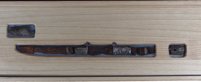 腐食した刀身と木製の柄に、銀製の装飾金具がはめ込まれた刀剣と、その両端の副葬品を、木箱に収め展示している銀装圭頭大刀の写真