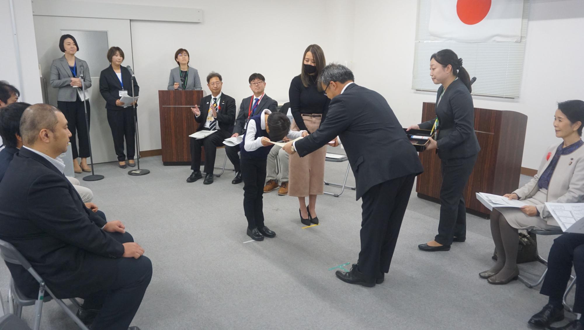 複数の関係者や参加者が見守っている中で、スーツを着た男性が入賞者の男の子に賞状を渡している様子を写した写真