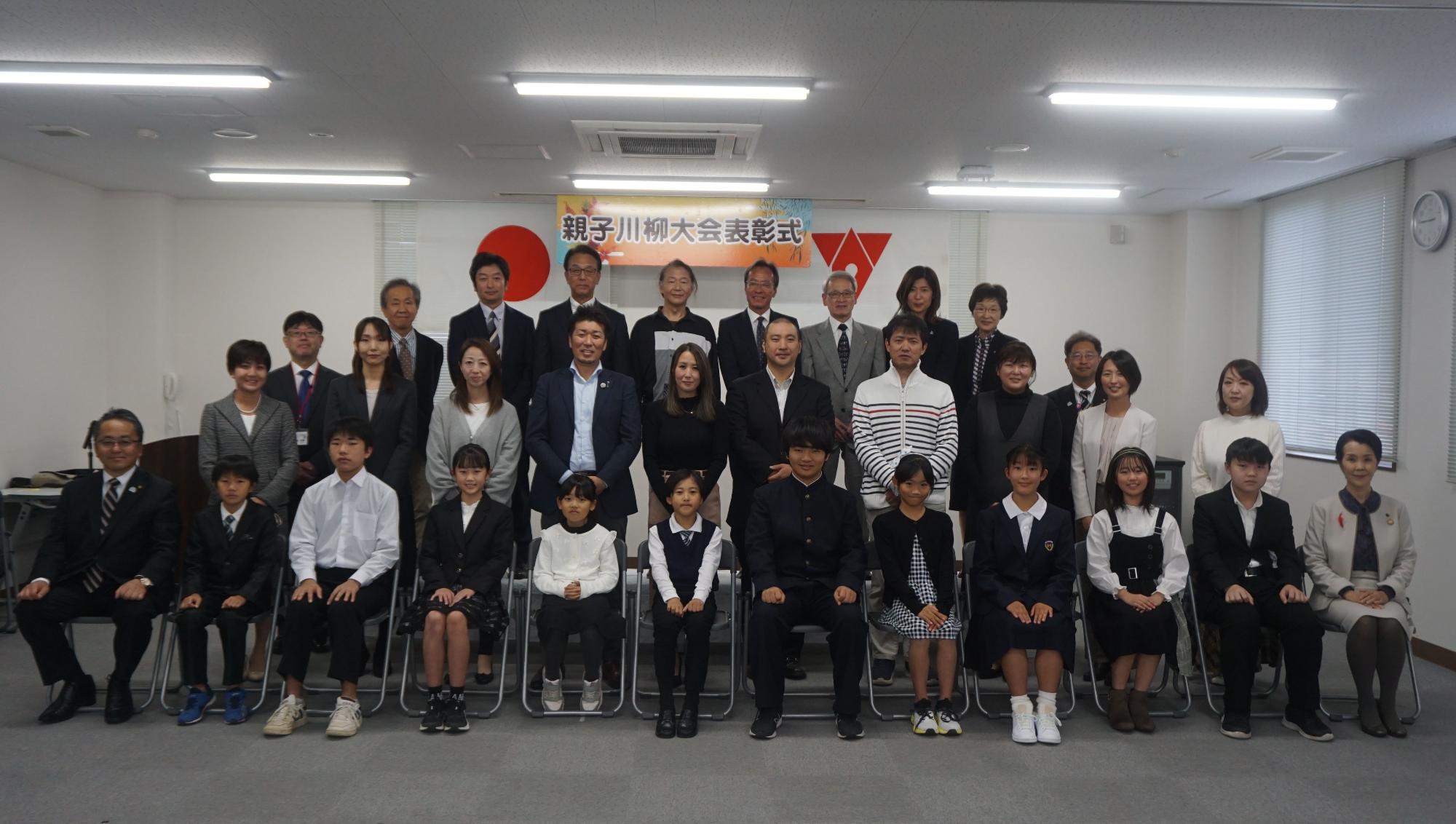 親子川柳大会表彰式と書かれた横断幕の前で、前列の椅子に入賞者の子どもたちが座り、その後ろに保護者や後援団体の方々が立ち3列に並んで写した記念写真