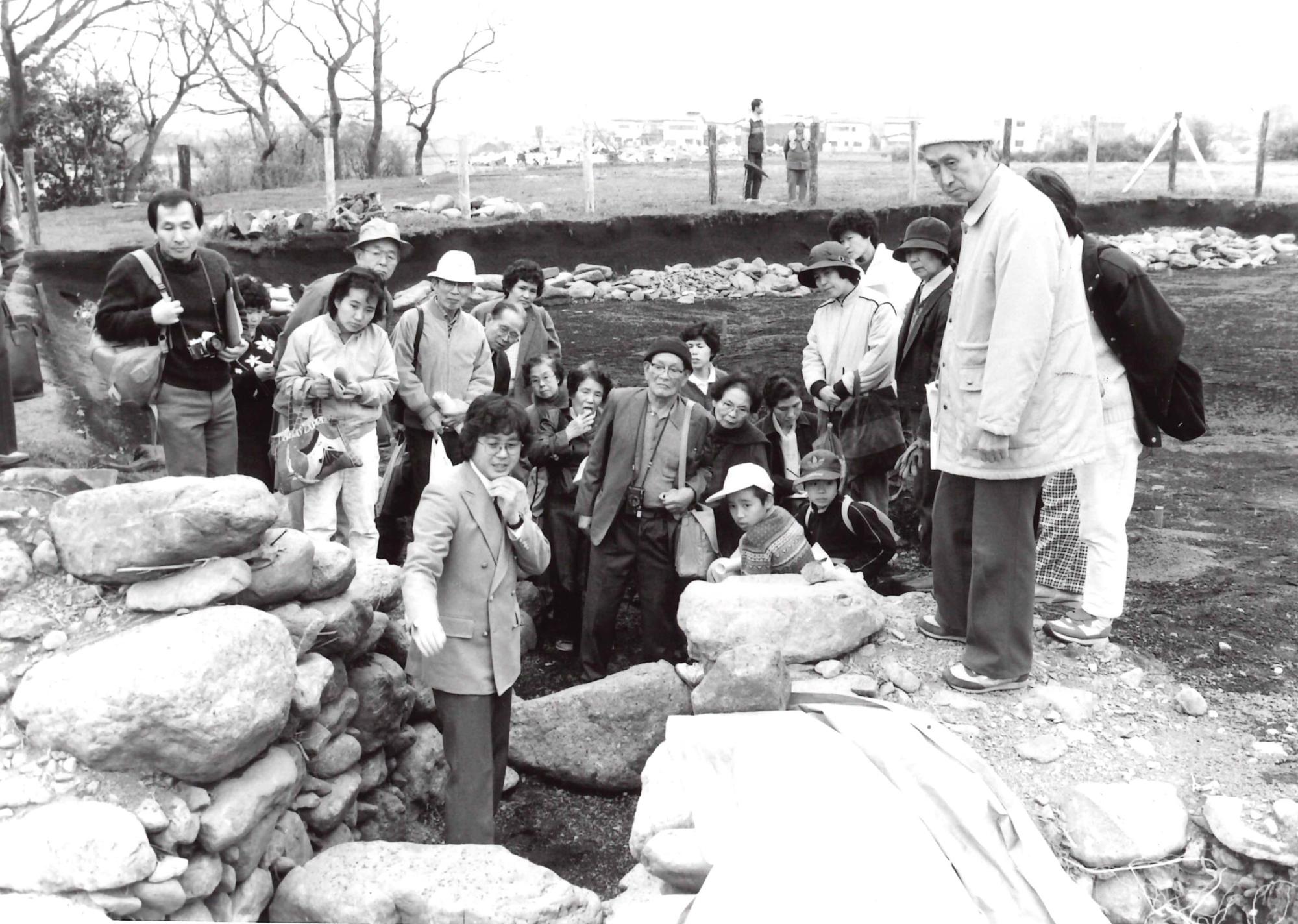 掘現場で、大勢の人々が大小様々な石が積み上げられた遺構を囲み、興味深そうに観察している様子のモノクロ写真