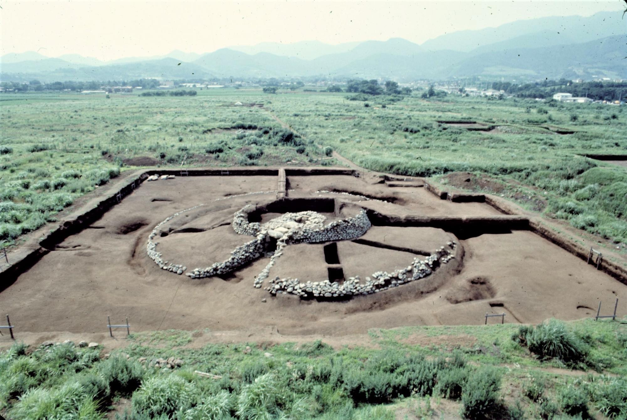 発掘された環状列石の遺構を上空から撮影した写真で、周囲は柵で囲まれており、遠景には山と田園風景が広がっている写真