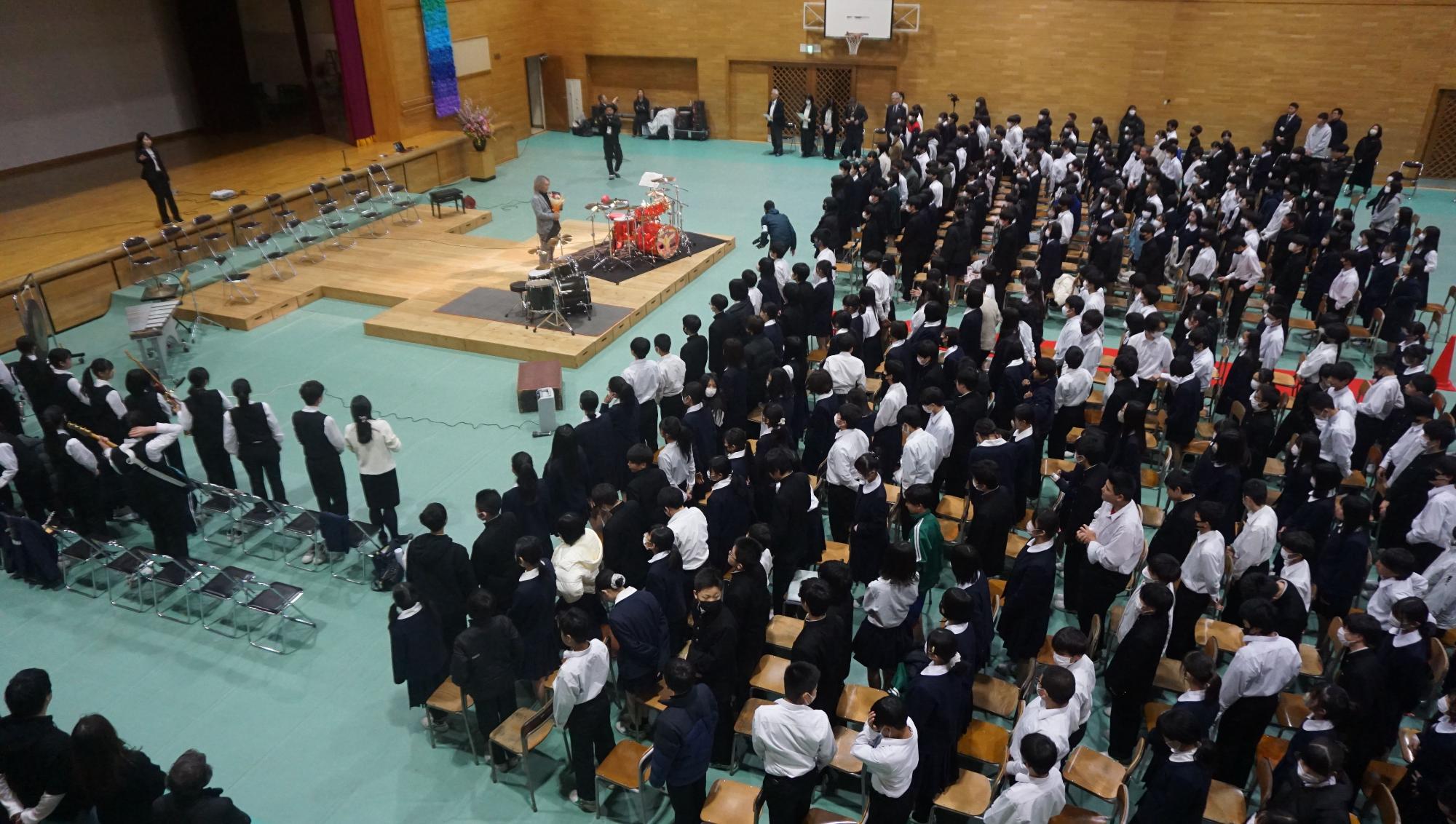 体育館の舞台前に設置されたステージにドラムセットが置かれ、その前にLUNA SEA 真矢さんが立っており、ステージの前や横に並んだ椅子の前に大勢の生徒たちが立っている講演の様子の写真
