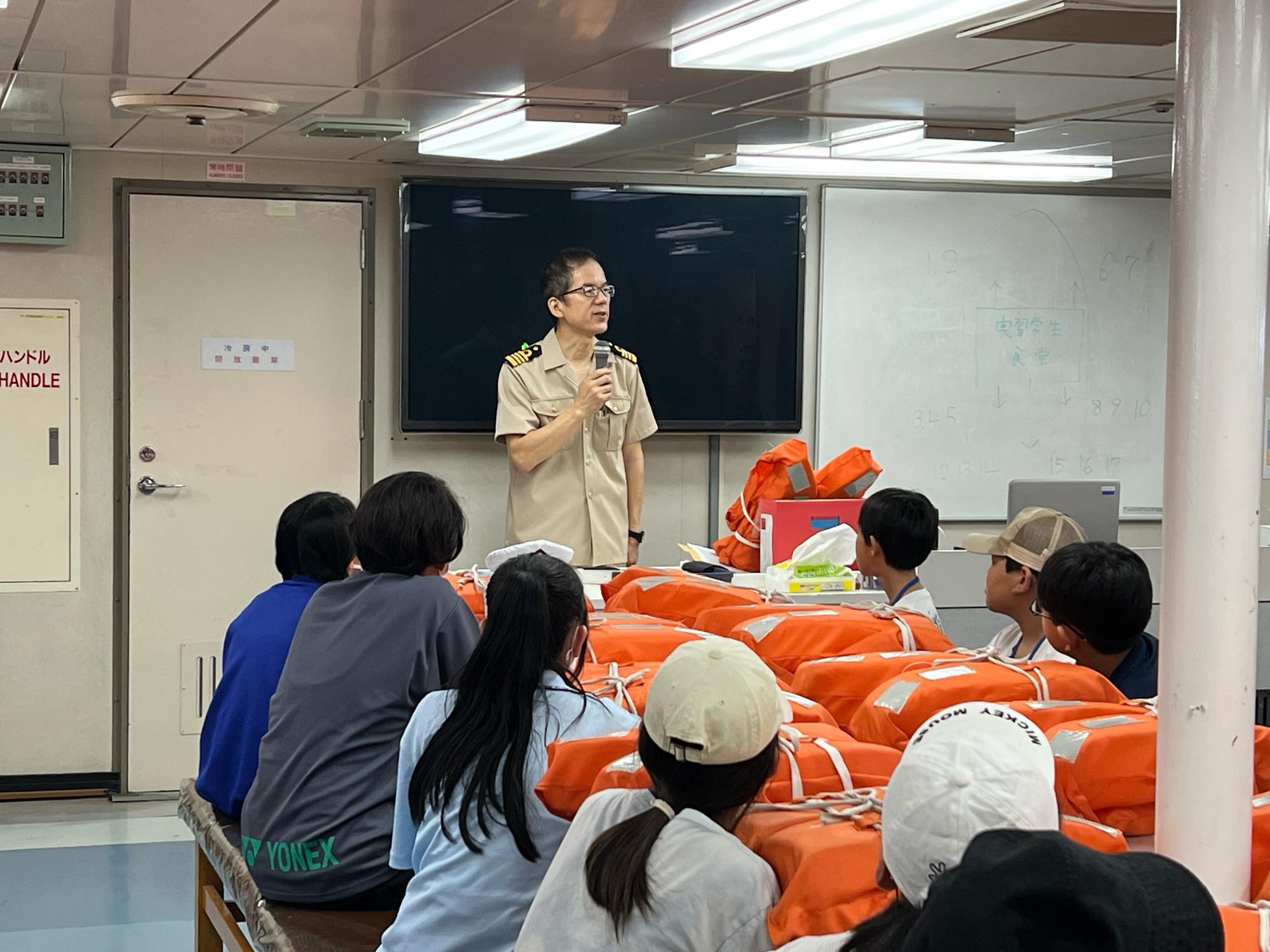 船内の一室で、制服を着た船長がマイクで話しており、複数の子どもたちがオレンジ色のライフジャケットが置かれたテーブルを囲んで座り、熱心に説明を聞いている様子の写真