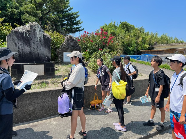 木々や草花を背景に建つ石碑の前で、数人の子供たちが引率者から話を聞いている様子の写真