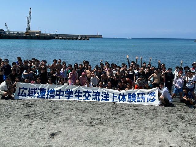 「広域連携中学生交流洋上体験研修」と書かれた横断幕を掲げ、海辺に集まった大勢の子どもたちがカメラに向かってポーズを取っている集合写真