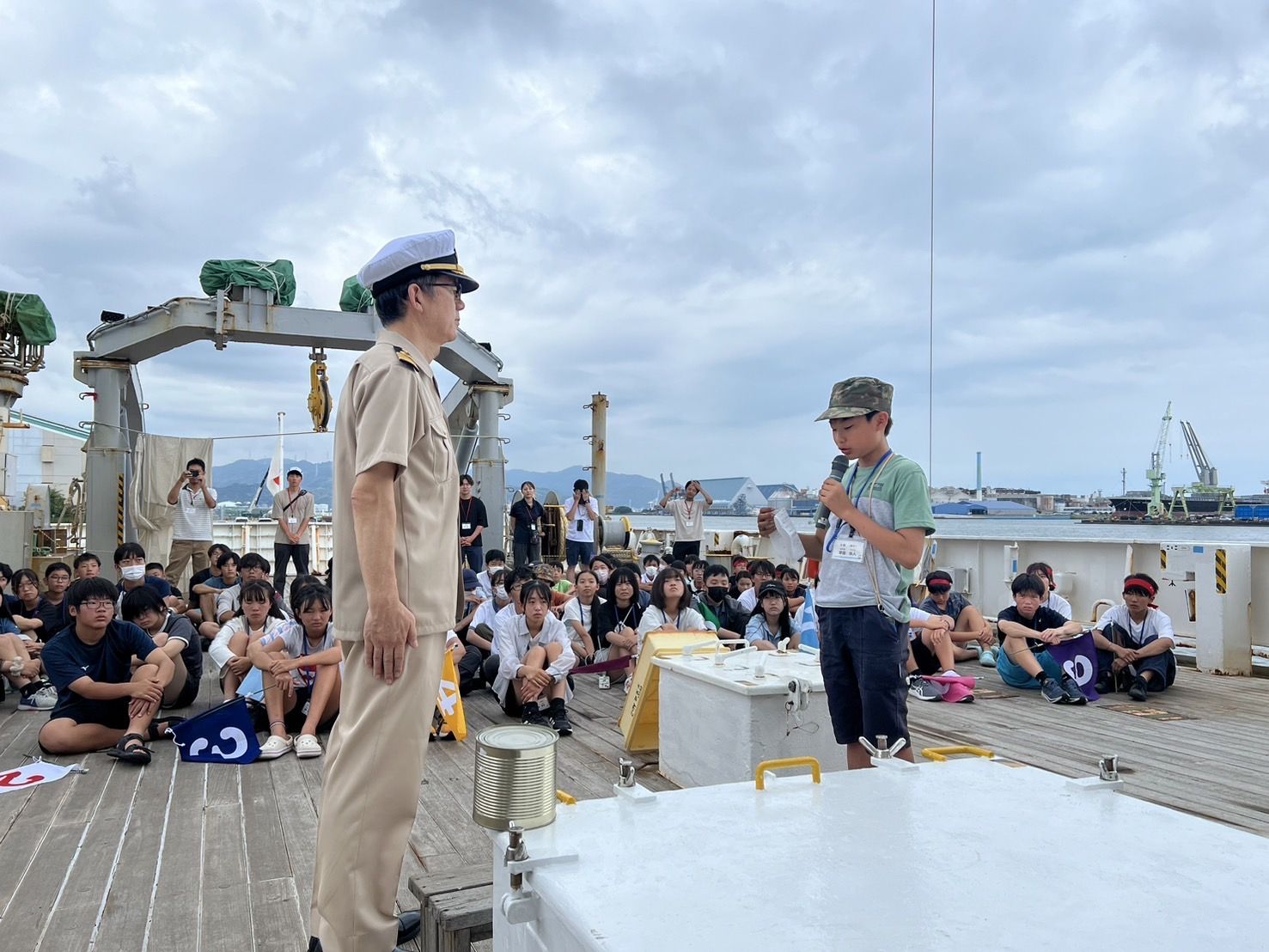 大勢の子どもたちが船のデッキに座り、制服姿の船長とマイクを持った少年が向かい合っている、帰港式の様子の写真