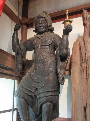 重厚な甲冑を身につけ、左手に鉾を持ち、右手に宝塔を掲げた勇ましい表情の木造の仏像を撮影した写真