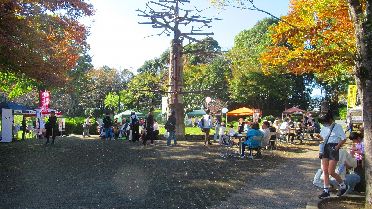 色づいた木々に囲まれた公園で、複数のテントが並ぶイベント会場に多くの人々が集い、賑わっている様子の写真