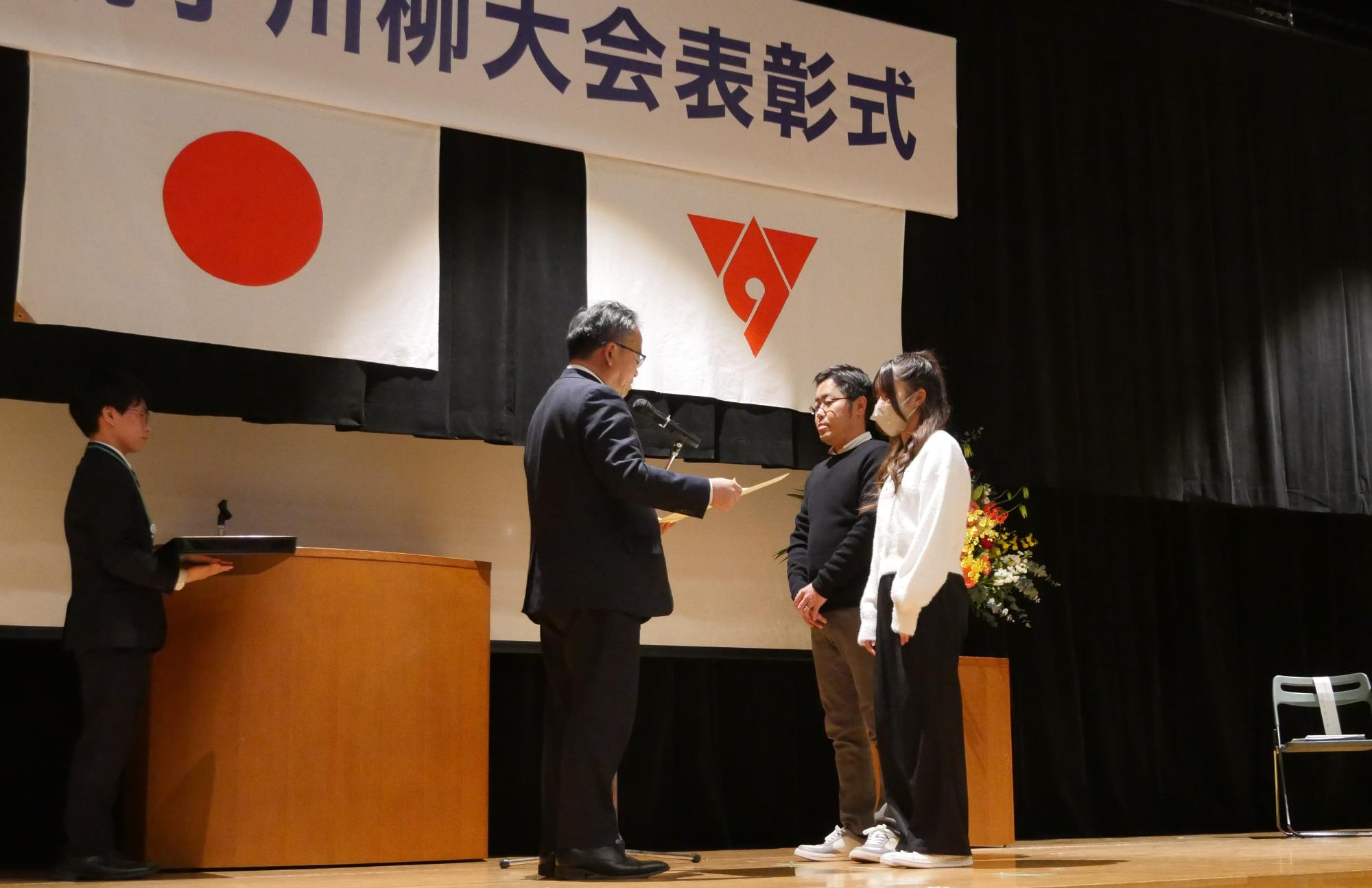 表彰式会場の舞台の演台の前で、2人の男女と向かい合った男性が、手に持った表彰状を読み上げている様子の写真