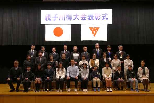 「親子川柳大会表彰式」の横断幕と日本国旗、秦野市の旗が掲げられた舞台の上で、受賞者、後援団体の方が前後3列に並んで写した記念写真