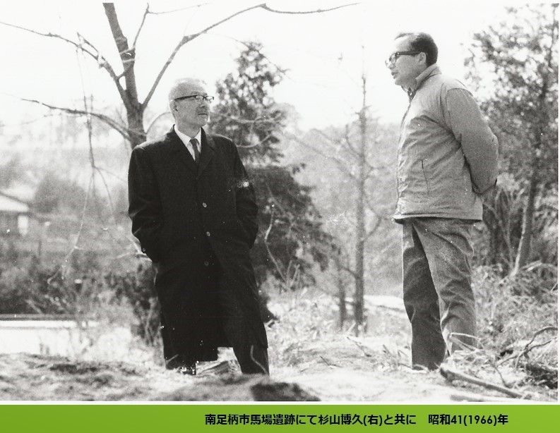 モノクロ写真に写るコートを着た直良信夫さんとジャンパーを着た杉山博久さんが、樹木の生い茂る屋外で会話する様子を捉えたモノクロ写真の下に「南足柄市馬場遺跡にて杉山博久（右）と共に 昭和41（1966）年」と添え書きがされた画像