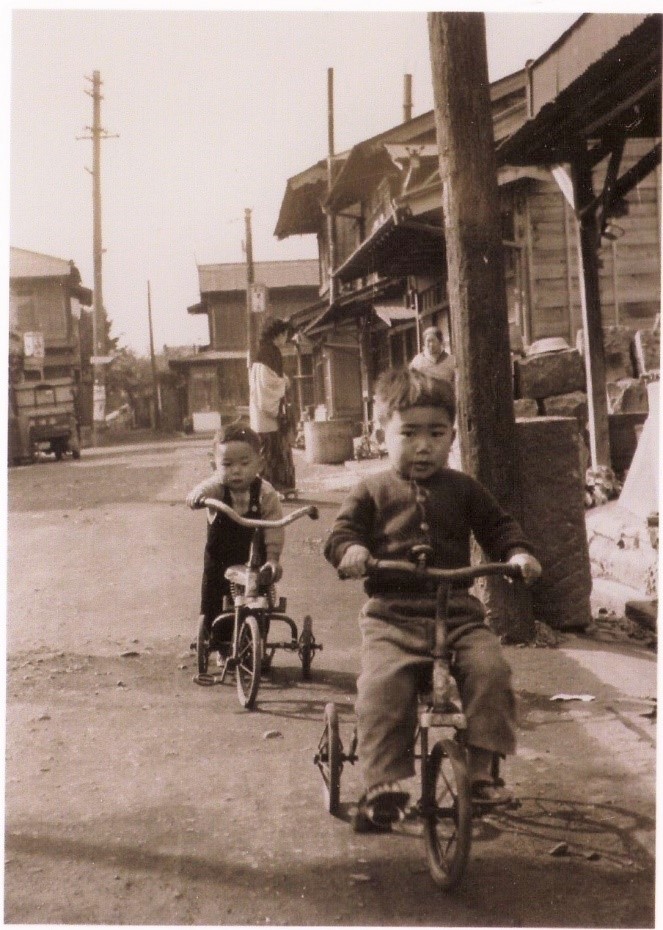木造の家屋が並ぶ中、二人の小さな男の子が三輪車に乗り、奥には和服の女性が立っている、昭和時代を思わせるモノクロ写真