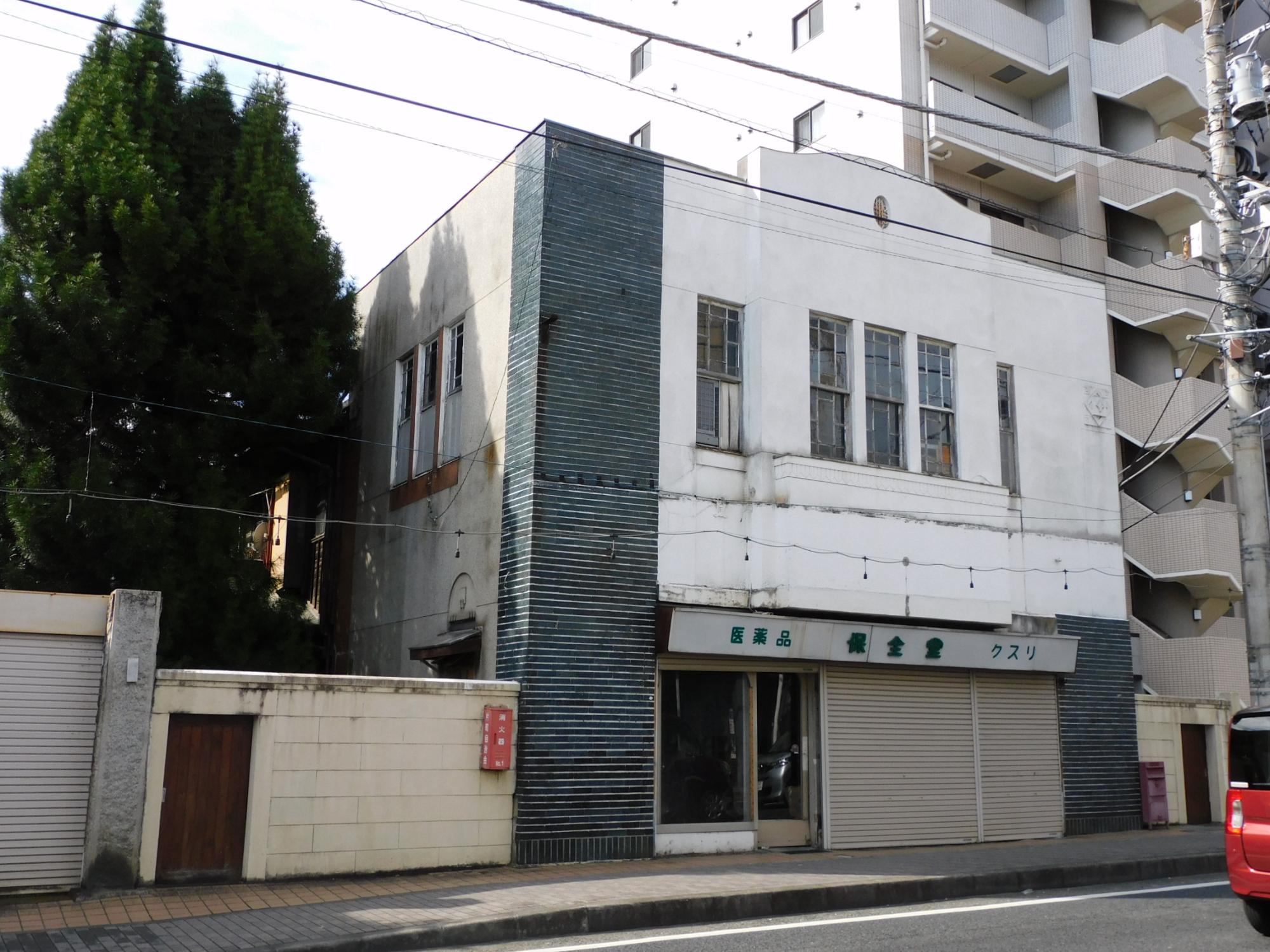 道路に面した歩道沿いに建っている、白壁で角に青系の幾何学模様のタイルレリーフの装飾がされた、1階がシャッターのついた薬局店舗となっている2階建ての建物の保全堂薬局店舗兼主屋の外観写真