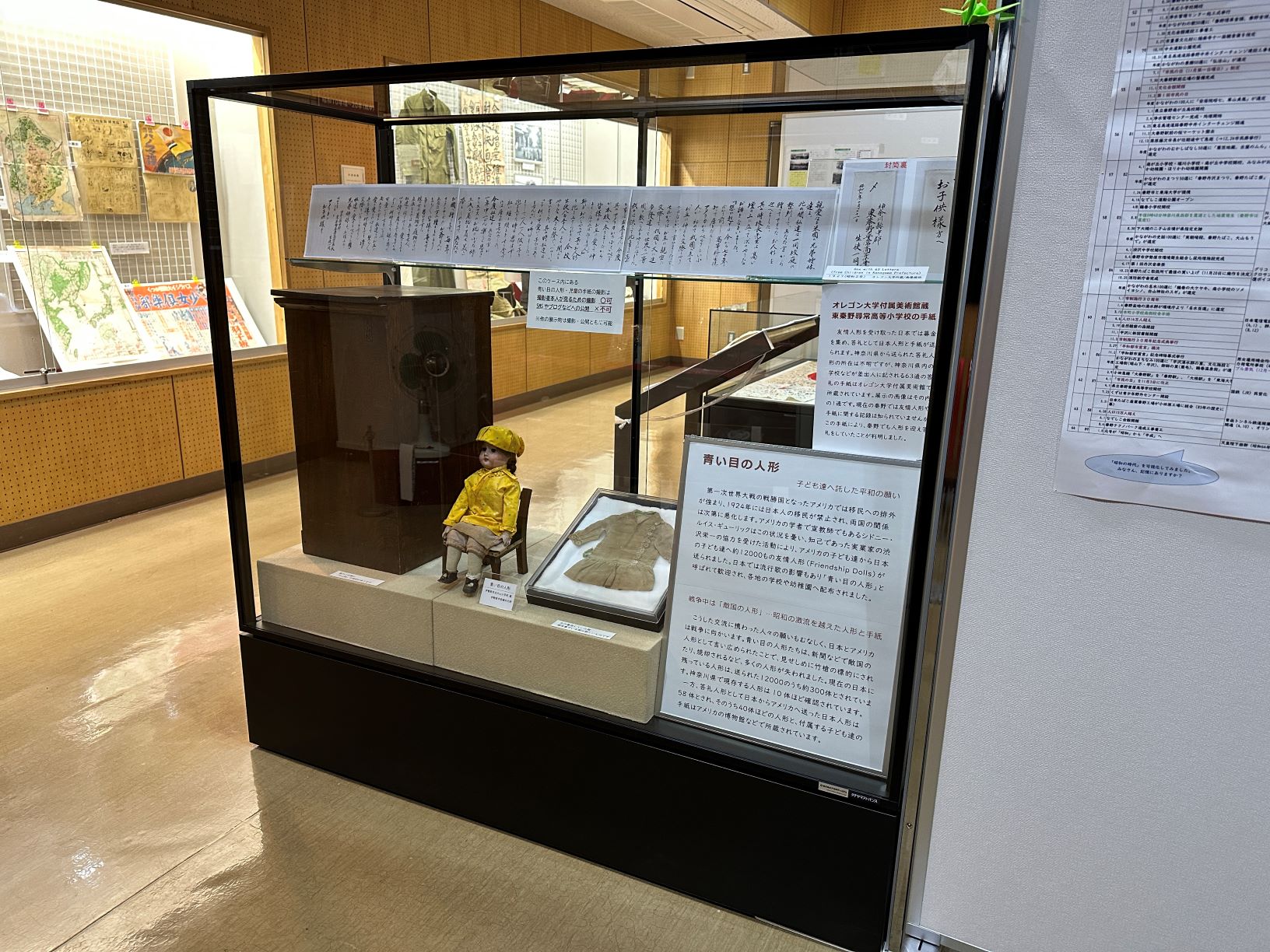 展示室の一角で、ガラスケースの中に座っている黄色い服と帽子を着た「青い目の人形」と、その傍らに展示されている人形の着物や解説パネルを写した写真