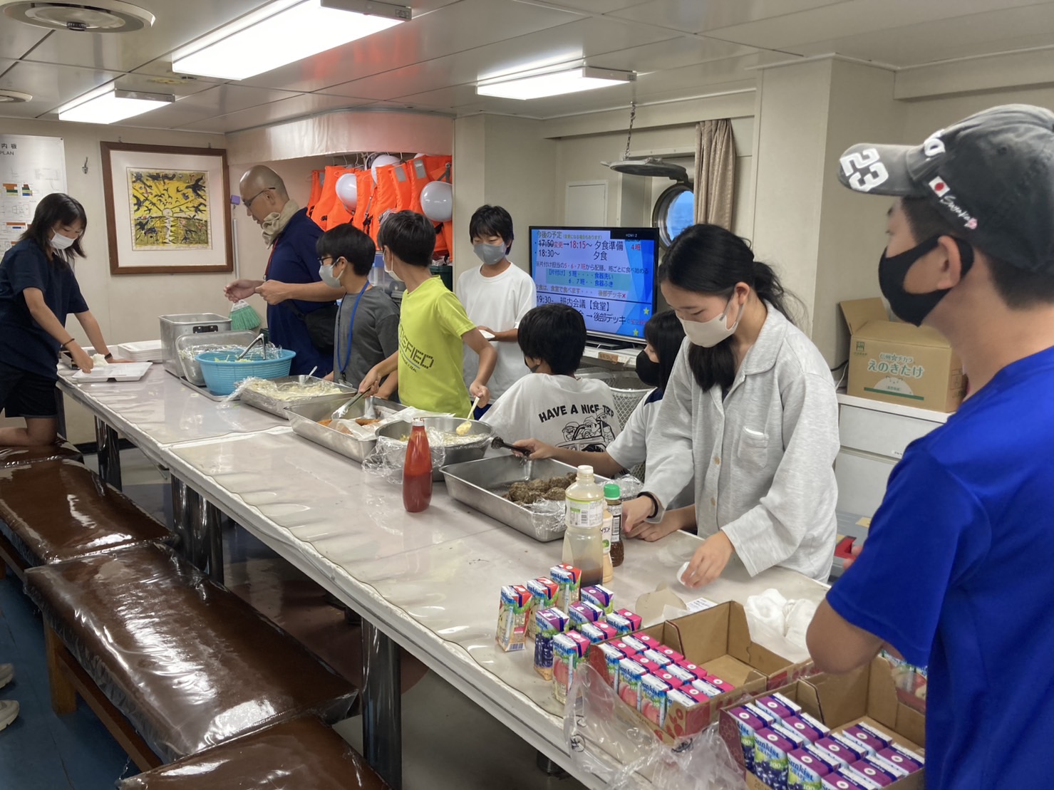 室内の長いテーブルで、マスクを着用している子どもたちと大人が食事の配膳をしている写真