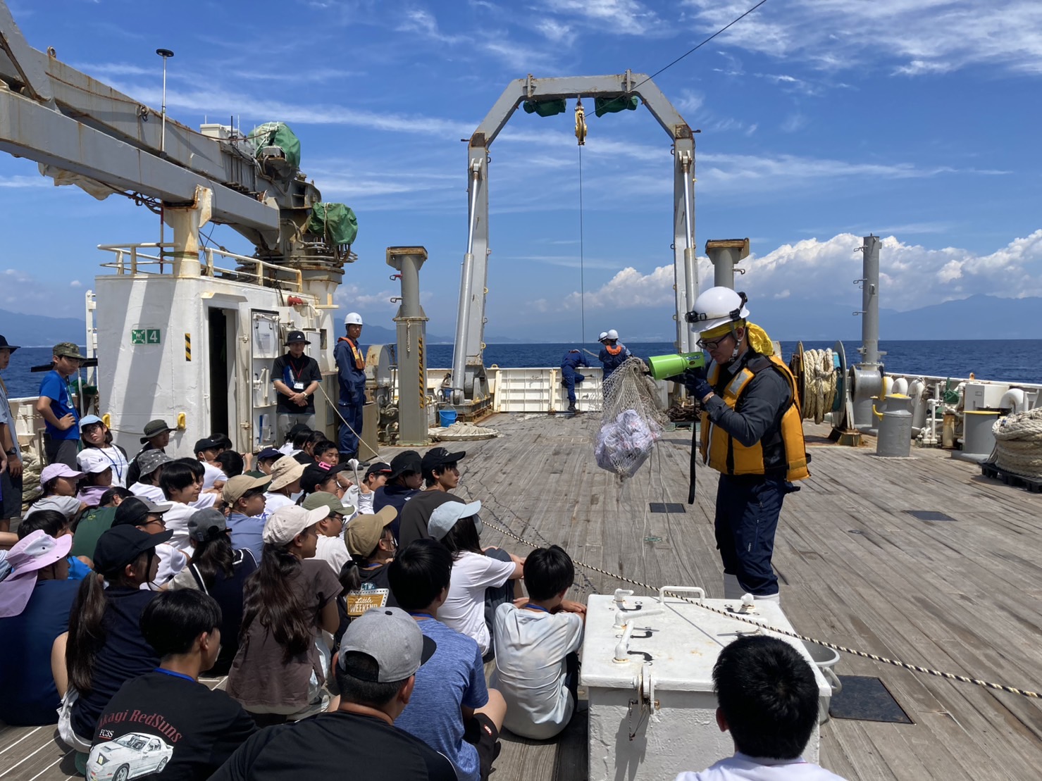 作業服とヘルメットとライフジャケットを着用した船員の男性が、前に集まって座った子どもたちに、網を見せながら説明している写真