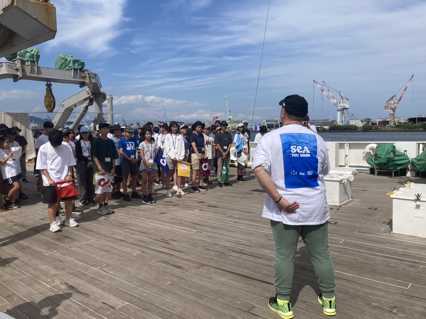 船のデッキに整列したたくさんの子どもたちの前に立った男性の後ろ姿の写真