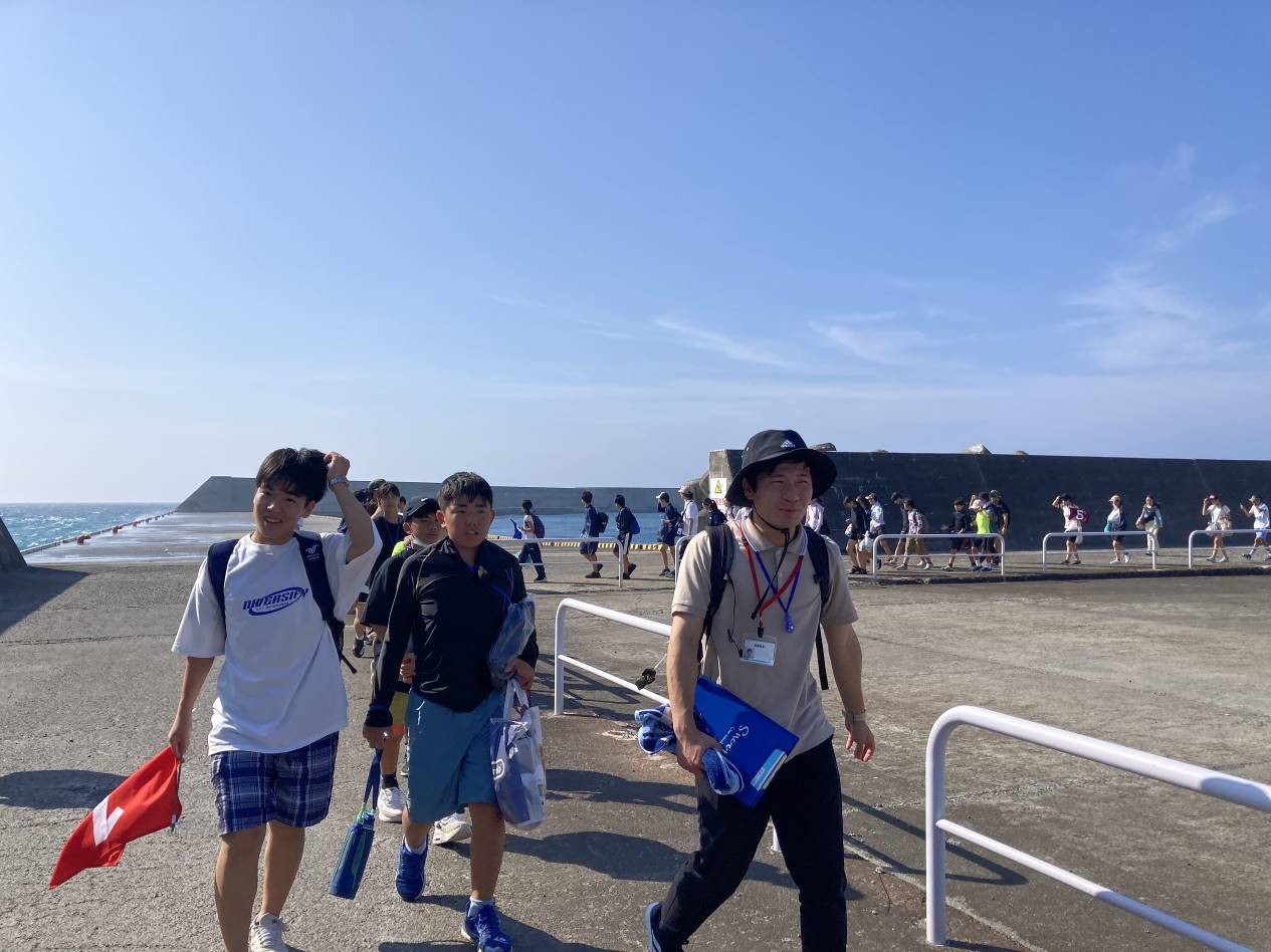 青空の下、港の海に面した広い歩道を大勢の子どもたちが引率者とともに歩いている様子の写真