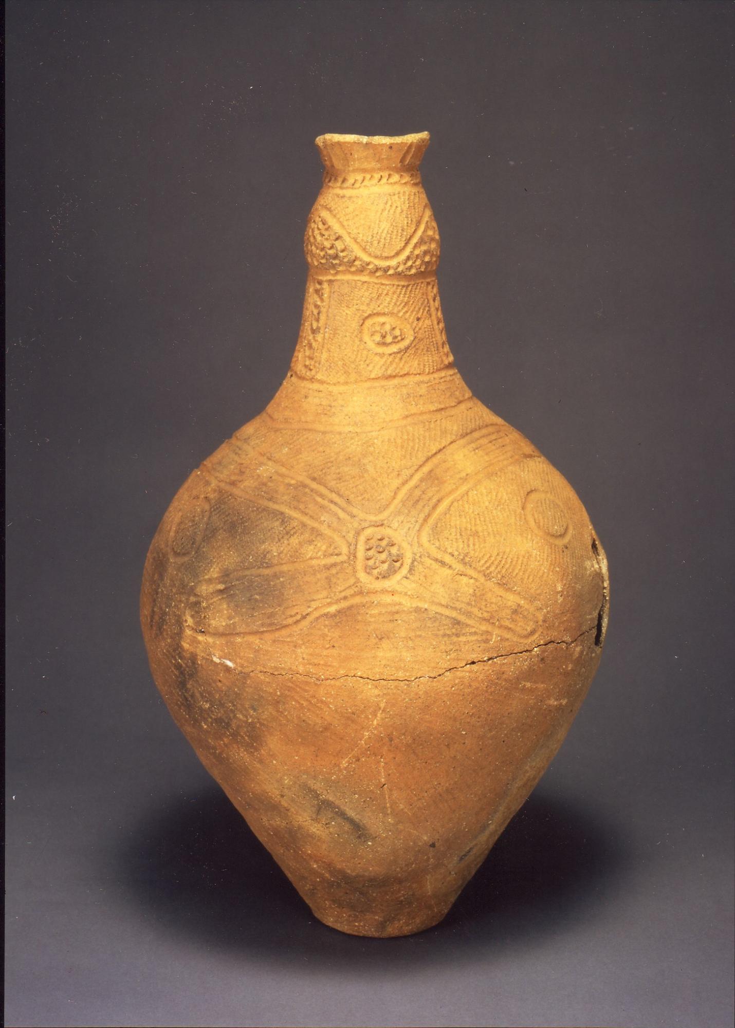 暗い背景に立つ茶褐色の土器で、細い首と丸い胴部を持ち、表面全体に縄目模様と幾何学文様が施され、胴部に水平な亀裂が見られる写真