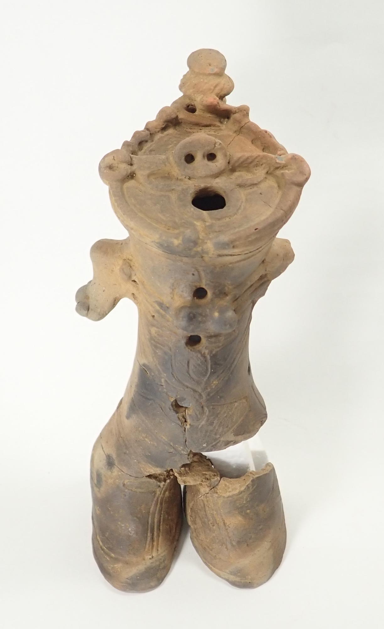 白い背景に、顔に豚のような鼻と口、頭部に小さな土器の破片のような装飾があり、胴体の股の部分が破損している人を模したような土偶の写真