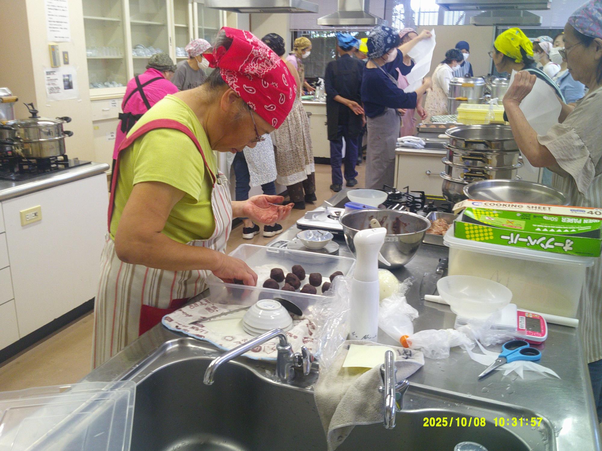 調理実習室で、赤いバンダナを着用した参加者が、シンク近くの作業台の上でトレイに入った茶色い丸い食材を扱っており、周囲には他の参加者や調理器具が見える様子の写真