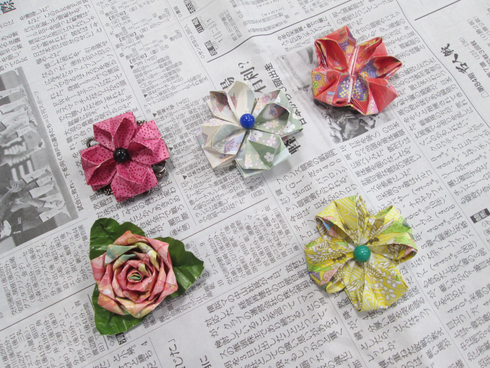 新聞紙の上に、赤、ピンク、白、黄色など色柄の異なる折り紙で作られた複数の立体的な花が配置されており、紙製のブローチ作品完成品が並んだ様子のクローズアップ写真