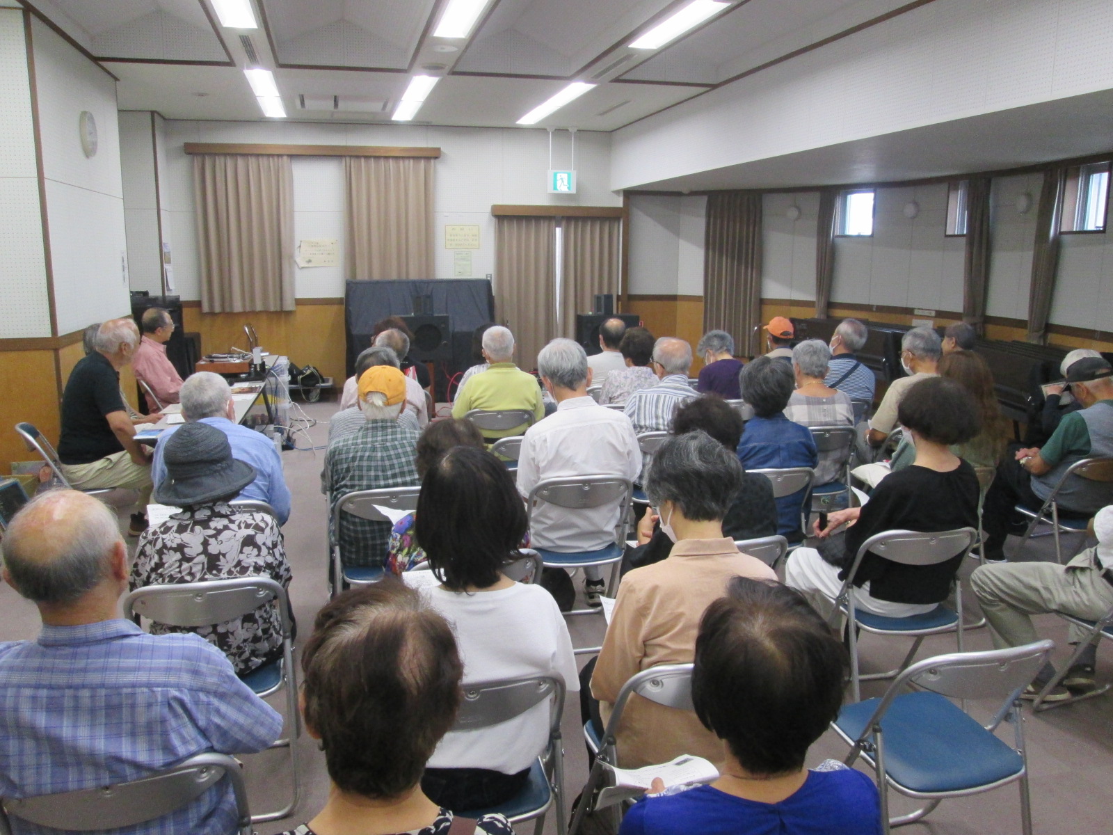 白い壁とベージュのカーテンのある室内で、多数の参加者が椅子に座り、前方の演台やスクリーンに向かって講演会や説明会に参加している様子の写真