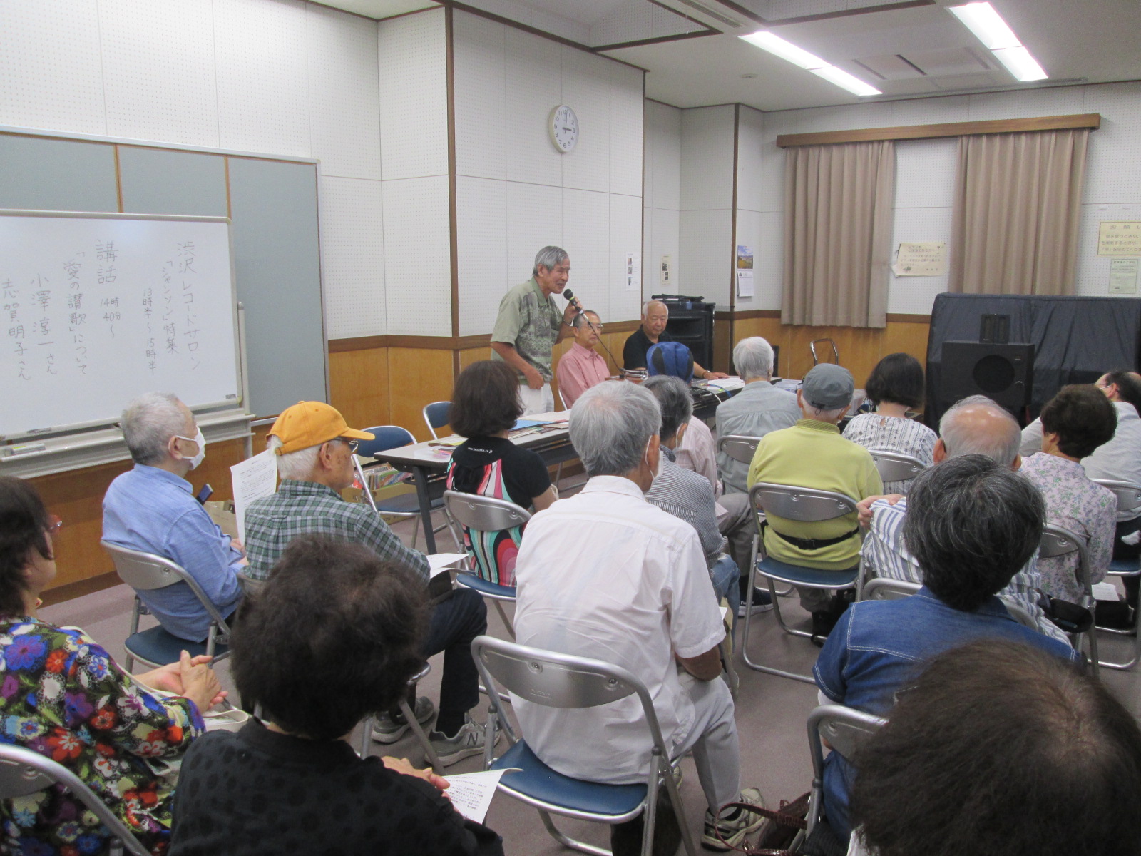 白い壁と木目調の腰壁がある室内で、多数の参加者が椅子に座り資料を手に持ちながら、マイクを持った講師の話を聞いている様子の写真