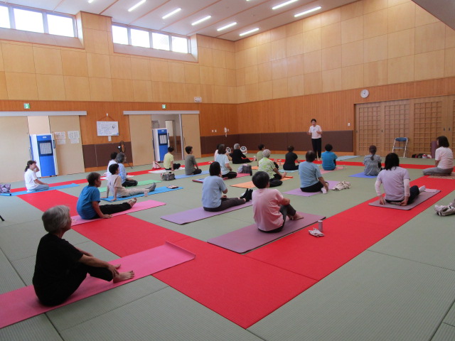 広々とした柔道場で、ヨガマットを敷いた多くの高齢者が中央の指導者の方を向いて座り、体操やヨガなどの運動を行っている様子の写真
