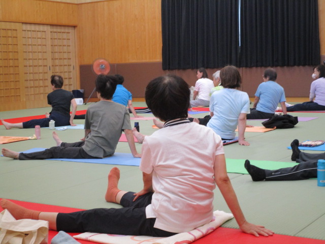 柔道場で、畳の上に敷かれたヨガマットの上で、体をひねるストレッチ運動を行っている大勢の人を背中の方から撮影した写真