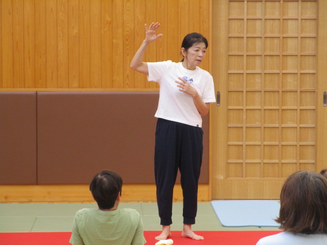 柔道場の室内で、白いTシャツと紺色のパンツ姿の女性が裸足で立ち、片手を上げて身振り手振りを交えながら参加者に何かを指導している様子の写真