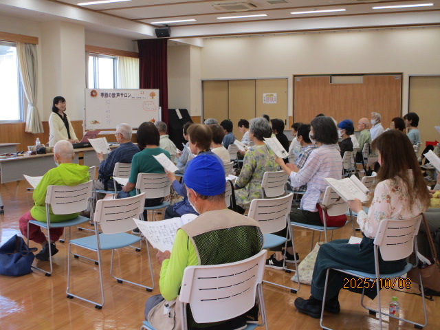 フローリングの広い室内で、前方の壇上には白いジャケットを着た指導者らしき女性が立ち、白い椅子の前で楽譜を持つ多くの高齢者が前を向いて座って発声練習している写真