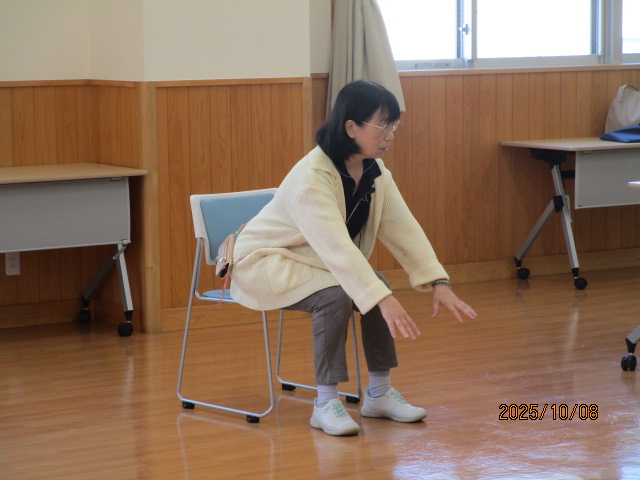 明るいフローリングの部屋で、カーディガンを着た女性が椅子に浅く腰掛けた状態で、手を前方に伸ばし、スクワットのような運動をしている様子を捉えた写真