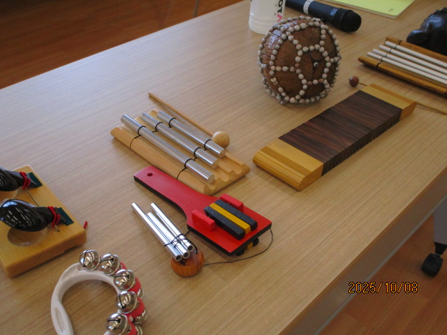 木のテーブルの上に、チャイムバー、タンバリン、マラカス、ギロなどの打楽器が並べられており、音楽イベントで使用される道具を上から撮影した写真