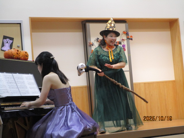 ピアノを紫色のロングドレスの女性が演奏しており、その横では緑色のドレスを着た魔女の仮装をした女性が杖を持ち、楽しそうに歌っている様子の写真