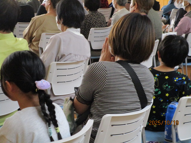 大勢の人が白いプラスチック製の椅子に座って前方を見ており、その中でボーダーラインのシャツを着た女性が手を合わせて座っている様子を、後方から捉えた写真