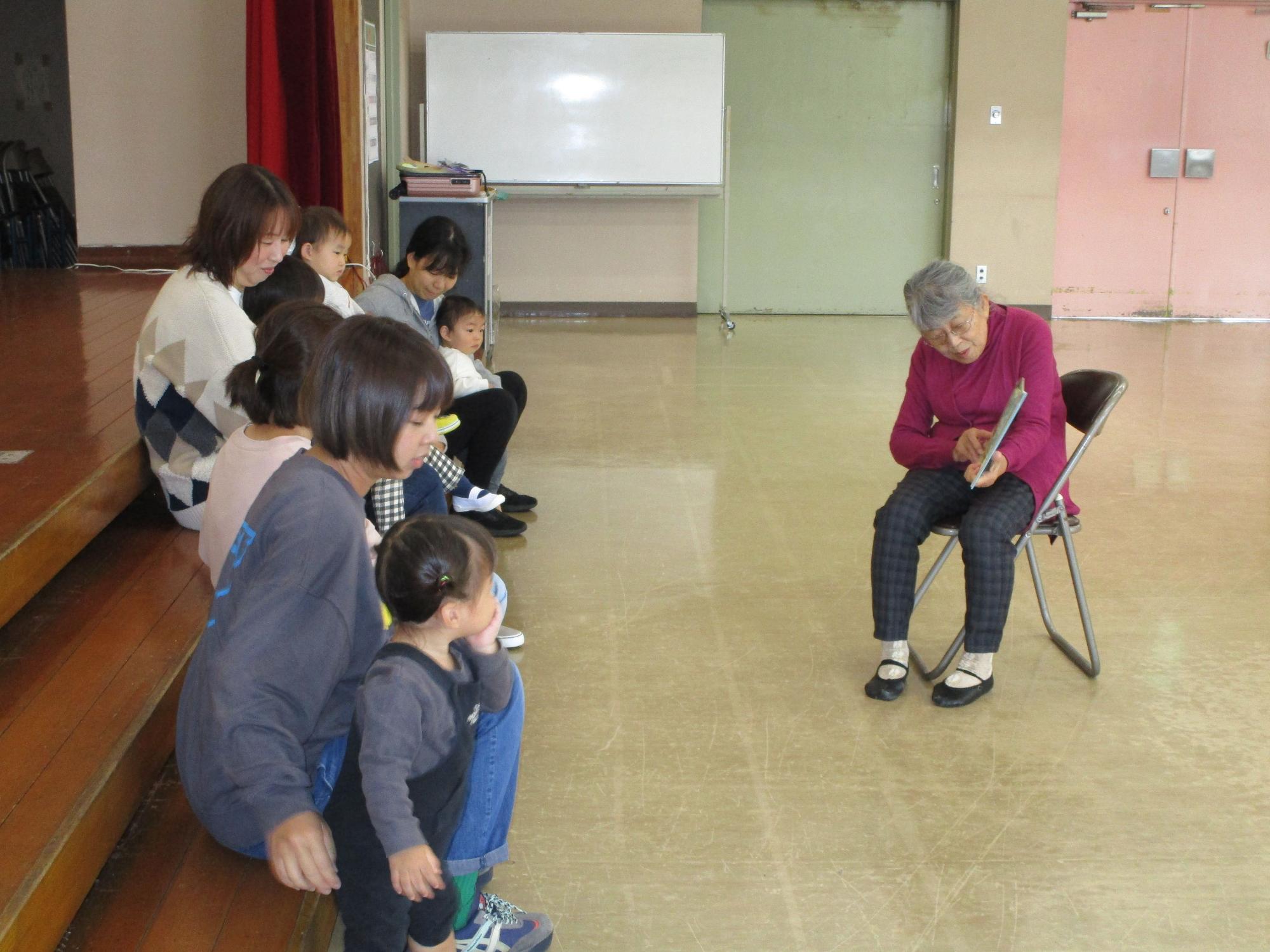 木製のステージに腰掛けている母親と幼い子どもたちと向かい合わせになり、一人の年配の女性が椅子に座って本を持ち、お話しをている写真