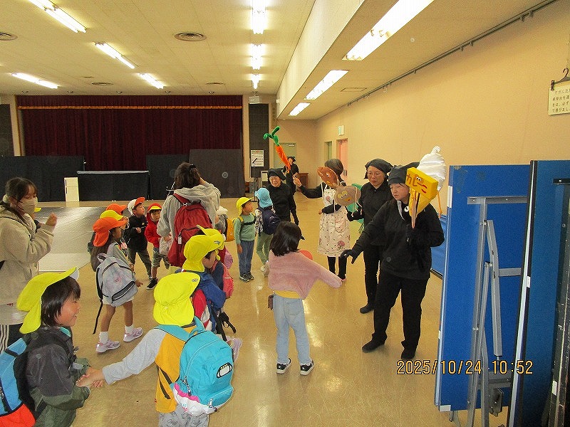 広い屋内スペースで、帽子をかぶった多くの子どもたちが、アイスクリームや人参、玉ねぎなどの食べ物の人形を持った方たちを近い距離で見ている写真