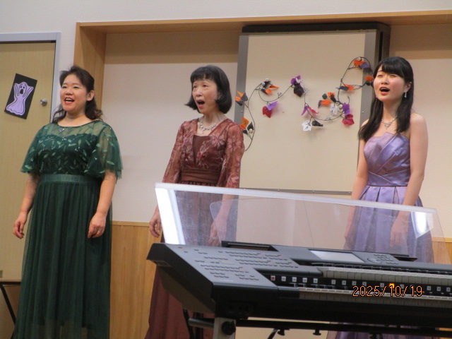 手前には電子キーボードがあるステージの上で、3人の女性がそれぞれ緑、茶、薄紫色のロングドレスを着て歌を披露している写真