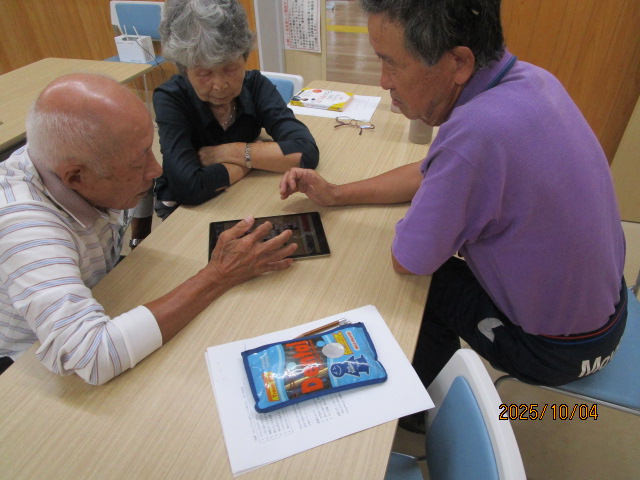 明るい会議室のような場所で、3人の高齢者がテーブルを囲んでおり、男性2人が中央に置かれたタブレット端末で囲碁をしているところを女性が眺めている写真