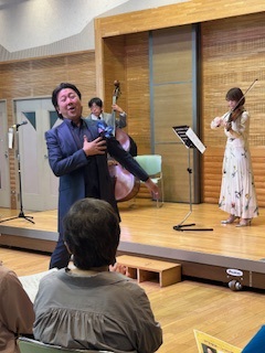 木目調のステージの前で、濃い青のスーツを着た男性ボーカリストが右手を胸に当てて歌い、後方にはヴァイオリンを弾く白いロングドレスの女性とコントラバス奏者が立っているのを参加者が見ている様子の写真